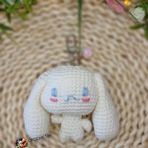 Pode incluir: Um chaveiro de crochê branco com um personagem de desenho animado com orelhas longas, olhos bordados azuis e bochechas rosadas. O chaveiro tem um fecho prateado e um pequeno pingente rosa. A palavra "CROCHET" é visível.