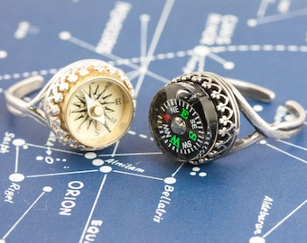 May include: Two silver rings with compass faces. One compass face is white with black markings and the other is black with green and white markings. The rings are on a blue background with white lines.
