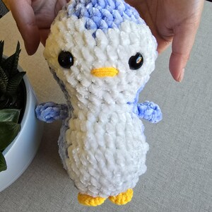 May include: A handmade plush penguin toy with a blue and white body, black eyes, and a yellow beak and feet. The penguin is made of soft, textured yarn and has small blue wings. The toy is held by two hands.
