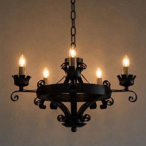 May include: A black metal chandelier featuring a circular frame with five upward-facing candle-style light sockets. The fixture is suspended by a matching black chain and displays decorative scrollwork accents at each light base and the centre.