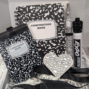May include: A black and white composition book themed stationery set including a spiral notebook, a vertical ID badge holder, a pen, and a heart-shaped charm, all featuring rhinestone accents. An Expo low odor dry erase marker is also included.