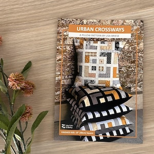 May include: A stack of four pillows with a pillow pattern titled "Urban Crossways" by Lisa Briese. The pillows have geometric designs in black, gray, and gold. The finished size is 19 inches (48cm) square.