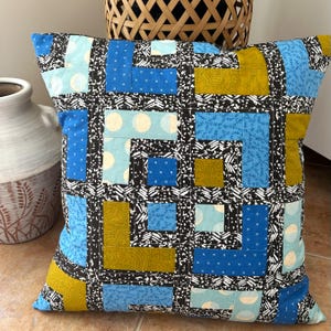 Könnte beinhalten: Quadratisches Zierkissen mit Patchwork-Design. Das Kissen hat ein geometrisches Muster mit blauen, gelben und weißen Stoffquadraten auf einem schwarz-weißen Hintergrund. Das Kissen hat eine quadratische Form.