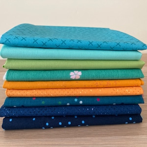 Peut inclure: Une pile de carrés de tissu colorés dans des tons de bleu, vert et orange. La couche supérieure est un tissu turquoise avec un motif de croix. D'autres tissus incluent des couleurs unies et des motifs à pois. Une fleur rose est visible sur une couche.