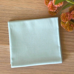 Könnte beinhalten: Ein gefaltetes, hellgrünes Stoffquadrat. Der Stoff hat eine glatte Textur und ist sauber gefaltet. Der Hintergrund ist eine hellbraune Holzoberfläche mit ein paar orangefarbenen und rosa Blüten in der oberen rechten Ecke.