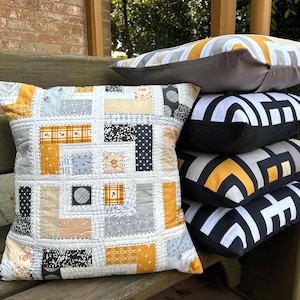 May include: A stack of five square throw pillows. The front pillow features a patchwork design with yellow, grey, black, and white fabric squares. The four pillows in the stack behind it display a geometric pattern in black, white, and yellow.