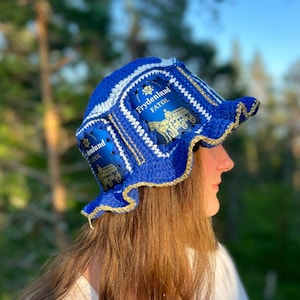 Peut inclure: Un chapeau bleu crocheté à la main avec des bordures blanches et dorées. Le chapeau porte l'inscription "Frydenlund FATØL" sur une étiquette. Le chapeau a un large bord et un design décoratif unique.