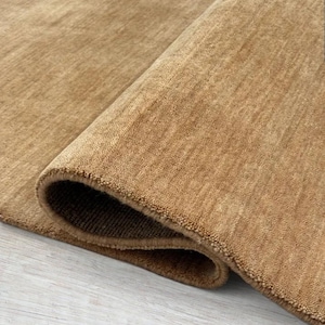 May include: A solid tan area rug with a dense, short-pile texture. The rug is partially folded over, revealing the thick, woven backing and the consistent, uniform color of the fibers across the entire surface of the floor covering.