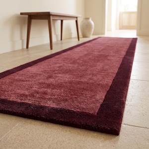 May include: A rectangular runner rug featuring a textured, dusty rose centre panel framed by a solid, dark burgundy border. The rug has a thick, plush pile and is laid out flat on a light-coloured stone floor, extending lengthwise into the room.