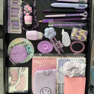 May include: A selection of purple stationery and beauty products displayed in a black tray. Items include pens, highlighters, a scrunchie, a notebook, eye patches, hand cream, and a small bag with smiley faces.