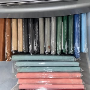 May include: A selection of colourful notebooks and journals, wrapped in plastic, in shades of brown, beige, black, grey, green, blue, and pink. The notebooks are stacked in a clear plastic container.
