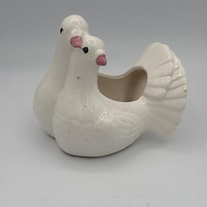 May include: A white ceramic dove planter with two doves. The doves have pink beaks and black eyes. The planter has a hollowed-out area for plants or decorations. The dove's tail feathers are detailed.