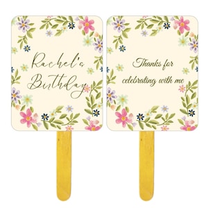 Könnte beinhalten: Zwei Partyfächer zum Geburtstag mit floralem Thema und Holzgriffen. Auf einem Fächer steht "Rachel's Birthday" und auf dem anderen "Thanks for celebrating with me". Die Fächer haben einen cremefarbenen Hintergrund mit einem floralen Kranzdesign.