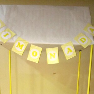 Puede incluir: Un puesto de limonada amarillo y blanco con una pancarta que dice "Lemonade". El puesto tiene una parte superior blanca y postes amarillos. La pancarta tiene letras amarillas sobre un fondo a rayas amarillas y blancas.