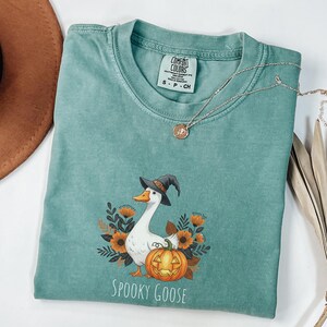May include: A teal Comfort Colors t-shirt with a Halloween design. It features a white goose in a witch's hat, a carved pumpkin, and the words "Spooky Goose". A silver necklace is draped across the shirt. The shirt is a small size.