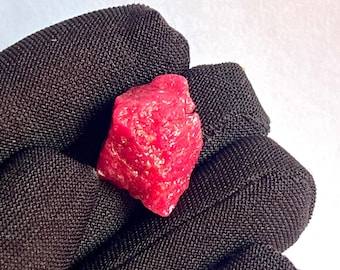 以下が含まれることがあります： 鮮やかな赤色の、原石のルビーの宝石。 ルビーは、粗く、質感のある表面と不規則な形状をしています。 宝石は暗い背景に表示されています。