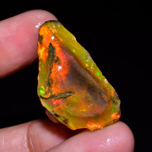 May include: A vibrant, raw opal gemstone with a mix of colors. The stone displays a fiery orange and yellow hue, with hints of green and brown. The opal is held in a hand against a black background.