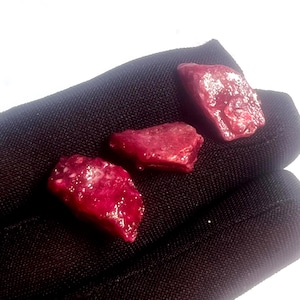Puede incluir: Tres piedras preciosas de rubí rojo oscuro en bruto. Las piedras preciosas tienen una superficie texturizada y se muestran sobre un fondo de tela negra. Los rubíes tienen forma irregular y parecen ser piedras naturales en bruto.