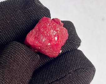 以下が含まれることがあります： 鮮やかな赤色の原石、未研磨のルビーの宝石。 粗い石は、テクスチャ加工された表面と不規則な形状をしています。 ルビーは黒い布で保持されており、石の色と対照的です。
