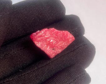 以下が含まれることがあります： 結晶構造を持つ、ラフカットの鮮やかな赤い宝石。ルビー色の石は黒い布の背景に置かれ、その自然な質感と色を際立たせています。宝石は不規則な形をしています。
