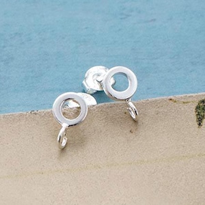 Peut inclure: Une paire de boucles d'oreilles à clous argentées. Chaque boucle d'oreille présente un petit cercle ouvert avec une tige et un petit crochet. Les boucles d'oreilles sont présentées sur une surface beige clair sur fond bleu.