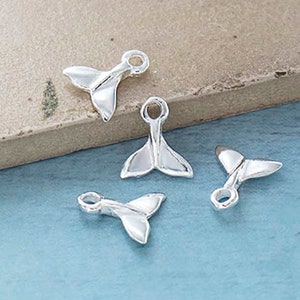 Puede incluir: Cuatro pequeños dijes plateados con forma de cola de ballena. Cada dije tiene un lazo para sujetarlo a una joya. Los dijes se muestran sobre una superficie azul claro con un borde texturizado marrón claro.