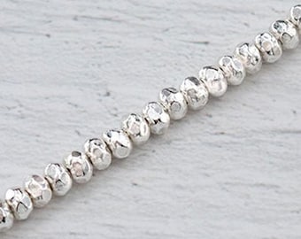 May include: A strand of small, faceted silver beads. The beads have a textured, irregular surface, reflecting light. The beads are strung together, creating a delicate, shimmering line. The background is a light, textured surface.