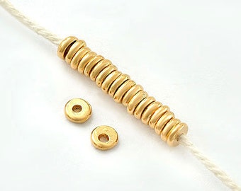 May include: Close-up of gold-colored jewelry beads and a string against a white background. The beads are small, round, and have a matte finish. Two loose beads are also visible. The string is a light beige color.