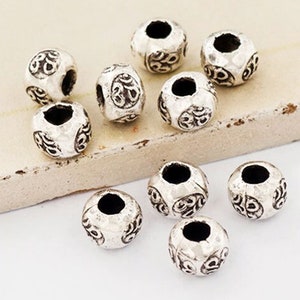 May include: A group of silver-toned metal beads with an intricate design. The beads are round and have a hole in the center for stringing.