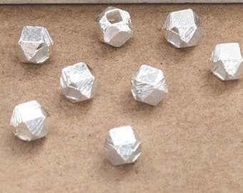 Può includere: Una collezione di dieci piccole perline sfaccettate color argento sparse su una superficie marrone. Le perline hanno una forma geometrica irregolare, che riflette la luce. Lo sfondo include un'area dipinta di bianco e azzurro.