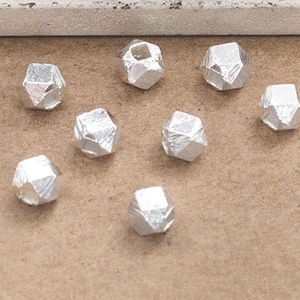 May include: Ten small, silver-coloured, faceted beads are scattered on a brown surface. The beads have an irregular, geometric shape, reflecting light. The background includes a white and light blue painted area.