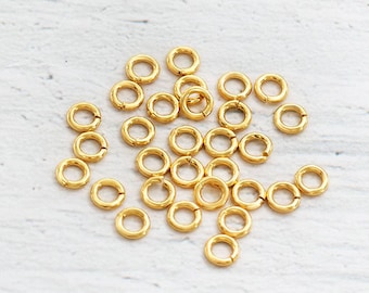 Può includere: Un mucchio di piccoli anelli di salto dorati. Gli anelli hanno un diametro di circa 5 mm e sono sparsi su una superficie bianca.