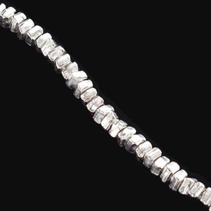 May include: A silver necklace with small, rectangular beads. The beads are strung together, creating a delicate and elegant design. The necklace is displayed against a black background, highlighting its metallic shine.