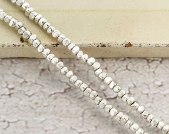 May include: Two strands of silver-colored beaded necklace. The beads are small, faceted, and evenly spaced, creating a delicate and shimmering effect. The necklace is displayed against a neutral, textured background, highlighting the jewelry's subtle shine.