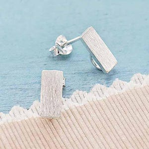 May include: A pair of silver earrings with a rectangular shape. The earrings have a textured surface and are designed to be worn as studs.