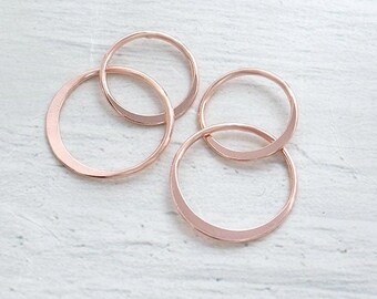 May include: Four rose gold-colored open circle charms are arranged on a white, textured surface. The charms are of varying sizes and are interlocked, creating a modern and minimalist design. The circles have a smooth, metallic finish.