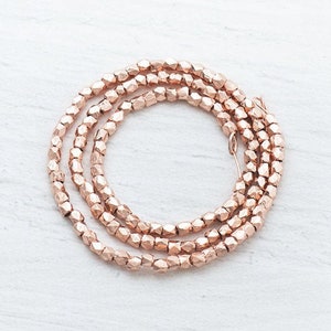 May include: A coiled strand of rose gold faceted beads, displayed on a white surface. The beads, with a metallic sheen, are geometric in shape, ideal for jewellery making or craft projects.