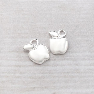 Puede incluir: Dos abalorios de manzana plateados con un detalle de hoja. Los abalorios son lisos y brillantes.