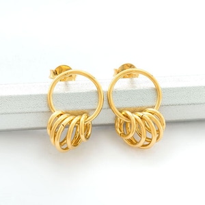 May include: A pair of gold-coloured stud earrings. Each earring has a circular design with a series of smaller, connected rings hanging below. The earrings are displayed on a white surface.