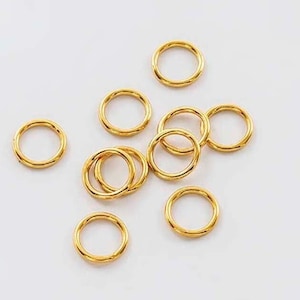 Puede incluir: Un grupo de diez anillos de salto dorados, también conocidos como anillos partidos, están esparcidos sobre una superficie blanca. Estos anillos se utilizan para conectar componentes de joyería.