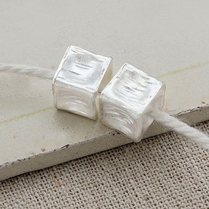 Puede incluir: Dos cuentas de cubo de plata con superficie texturizada enhebradas en un cordón blanco.