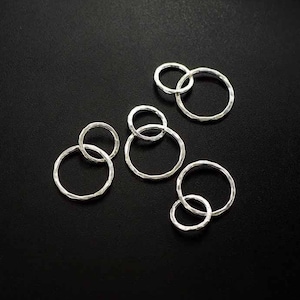 Può includere: Cinque anelli di salto in argento, ognuno con una superficie testurizzata, sono disposti su uno sfondo nero. Gli anelli sono di dimensioni diverse e sono collegati a coppie.