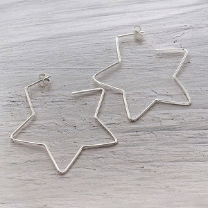 May include: A pair of silver star-shaped hoop earrings. The earrings are made of thin wire and have a simple, modern design.