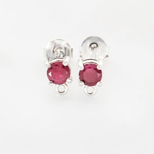 Puede incluir: Un par de pendientes de plata con tachuelas, cada uno con una piedra preciosa redonda de color rojo intenso. Los pendientes tienen un poste y un pequeño lazo en la parte inferior. Fondo blanco.