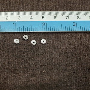 Puede incluir: Cuatro cuentas espaciadoras de metal de color plateado, cada una con un di&aacute;metro aproximado de 1,25 cm, est&aacute;n dispuestas sobre una superficie de tela marr&oacute;n. Una regla es visible en el fondo.