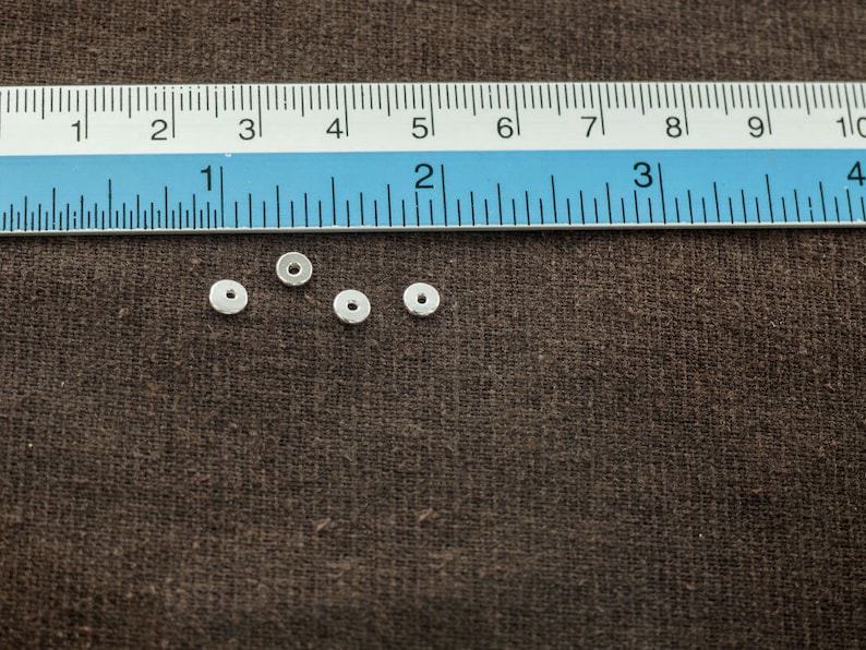 Puede incluir: Cuatro cuentas espaciadoras de metal de color plateado, cada una con un di&aacute;metro aproximado de 1,25 cm, est&aacute;n dispuestas sobre una superficie de tela marr&oacute;n. Una regla es visible en el fondo.