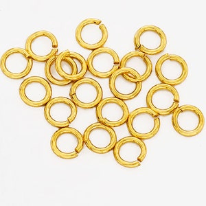 以下が含まれることがあります： 白い表面に、20個の金色のジャンプリングが散らばっています。ジャンプリングは、ジュエリーの金具の一種です。リングは小さく、滑らかで磨かれた仕上げになっています。
