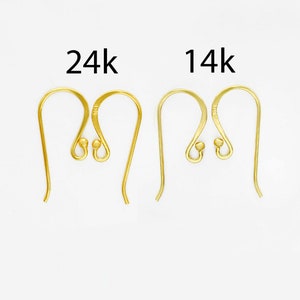 Op de afbeelding: Twee paar goudkleurige metalen oorhaken, waarbij het linkerpaar is gelabeld met 24k en het rechterpaar met 14k. Elke haak heeft een gebogen draadontwerp met een klein bolletje aan de basis voor het bevestigen van sieradenonderdelen.