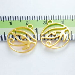 Puede incluir: Dos amuletos de metal dorado en forma de Ojo de Horus, cada uno mide aproximadamente 2,5 cm de di&aacute;metro. Los amuletos tienen un dise&ntilde;o recortado y se muestran contra una regla blanca.