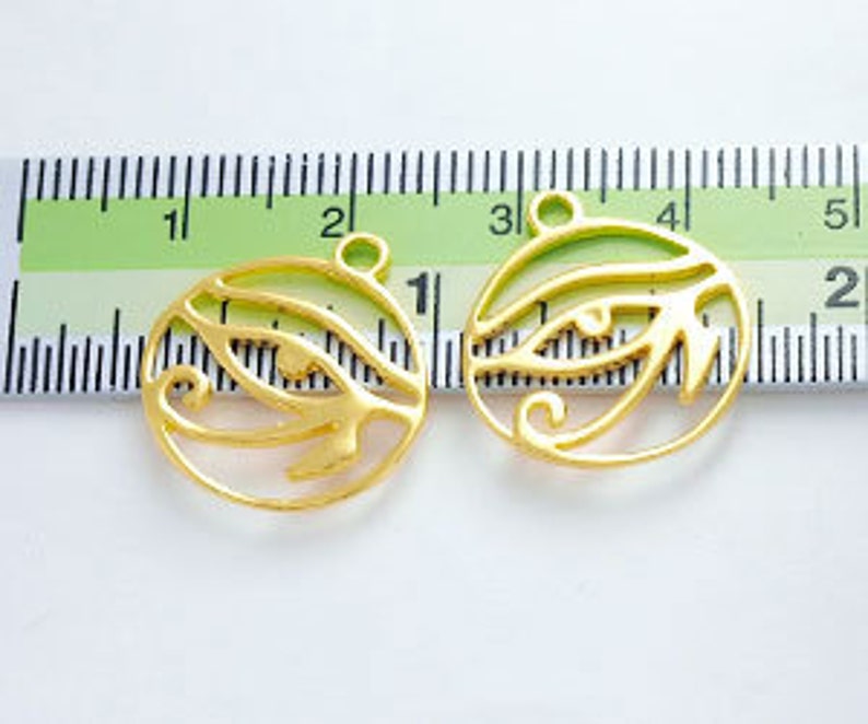 Puede incluir: Dos amuletos de metal dorado en forma de Ojo de Horus, cada uno mide aproximadamente 2,5 cm de di&aacute;metro. Los amuletos tienen un dise&ntilde;o recortado y se muestran contra una regla blanca.